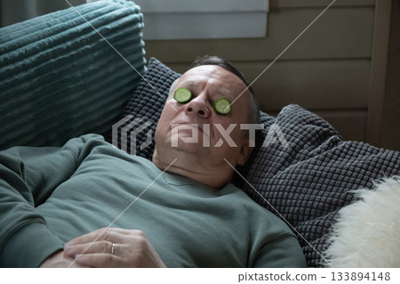 Resting caucasian man lying on sofa with cucumber slices on eyes, practicing simple home skincare and relaxation indoors. Home Leisure Activities Resting caucasian man lying on sofa with cucumber slices on eyes, practicing simple home skincare and relaxation indoors. Home Leisure Activities 133894148