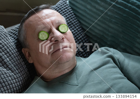Resting caucasian man lying on sofa with cucumber slices on eyes, practicing simple home skincare and relaxation indoors. Home Leisure Activities Resting caucasian man lying on sofa with cucumber slices on eyes, practicing simple home skincare and relaxation indoors. Home Leisure Activities 133894154