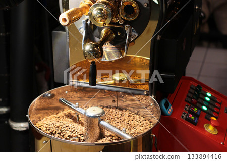 A modern coffee roasting machine filled with brown coffee beans stands next to a control panel with colorful buttons. The professional setup enables precise roasting and flavor control. 133894416