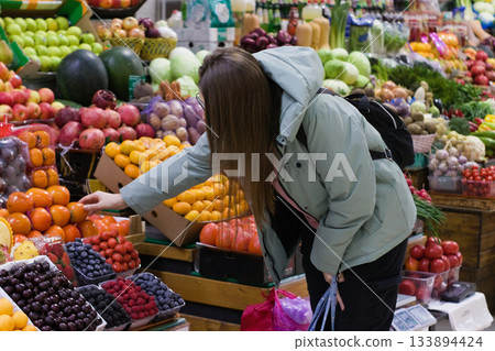 At a busy produce market, a young woman in a light green jacket reaches out to pick up fruit from a colorful selection. The scene is lively with a variety of fruits around her. At a busy produce market, a young woman in a light green jacket reaches out to pick up fruit from a colorful selection. The scene is lively with a variety of fruits around her. 133894424