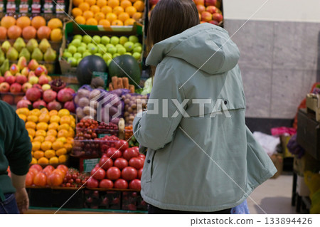 In a vibrant marketplace, a young woman with a blue shopping bag pauses in front of a display of fresh fruits and vegetables. Her attention is fixed on the colorful produce. In a vibrant marketplace, a young woman with a blue shopping bag pauses in front of a display of fresh fruits and vegetables. Her attention is fixed on the colorful produce. 133894426