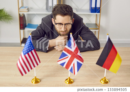 Pensive businessman sitting at desk looking at flags of different countries 133894505
