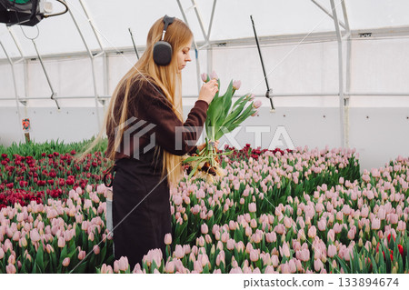 Woman collecting flowers peacefully, Young lady working with flowers calmly in greenhouse, Youthful female tending to flowers thoughtfully while immersed in music and nature ambiance 133894674