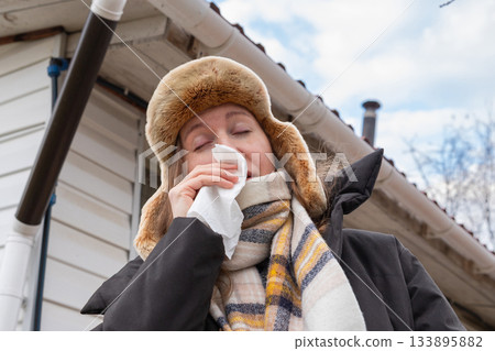 Caucasian woman wearing a winter hat is seriously ill and sneezing into a white paper handkerchief. She feels weak, unwell, and has a virus and chills all over her body, allergy 133895882