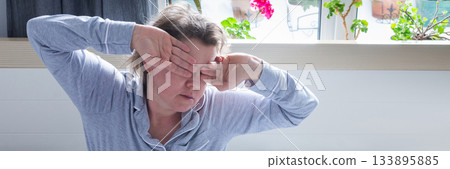 A 50-year-old woman appears tired and distressed as she sits up in bed. She rubs her eyes, indicating a lack of sleep, while surrounded by soft bedding in a bright room, banner A 50-year-old woman appears tired and distressed as she sits up in bed. She rubs her eyes, indicating a lack of sleep, while surrounded by soft bedding in a bright room, banner 133895885