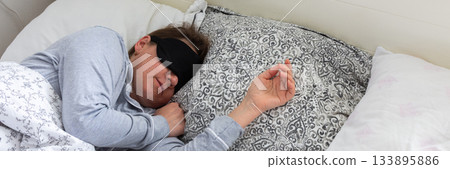A 50-year-old woman is peacefully sleeping in a cozy bed. She wears a sleep mask and is snuggled under soft blankets, creating a serene atmosphere for rest, banner A 50-year-old woman is peacefully sleeping in a cozy bed. She wears a sleep mask and is snuggled under soft blankets, creating a serene atmosphere for rest, banner 133895886