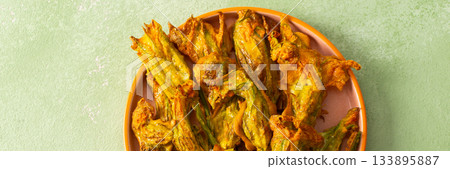 Golden-brown deep-fried zucchini flowers are arranged on a vibrant plate. The dish is set on a floral-patterned napkin, enhancing its appetizing appearance, banner Golden-brown deep-fried zucchini flowers are arranged on a vibrant plate. The dish is set on a floral-patterned napkin, enhancing its appetizing appearance, banner 133895887