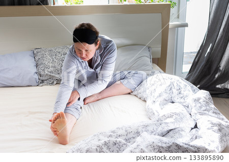 In a serene bedroom setting, a 50-year-old woman stretches her legs while sitting in bed. Sunlight pours in through the window, creating a peaceful atmosphere for a refreshing morning routine 133895890