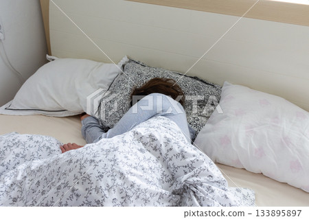 A middle-aged woman is sleeping soundly in her bed, wearing a sleep mask. She is surrounded by soft pillows and a warm blanket, taking a break from her daily routine 133895897