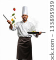 Happy Professional Male Chef Tossing Fresh Vegetables with Spatula and Frying Pan Against White Background. 133895939