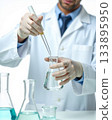 Close-up of Male Scientist in Lab Coat Pipetting Clear Liquid into Erlenmeyer Flask in Studio. 133895950