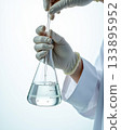 Gloved Hands of Scientist Adjusting Pipette in Large Erlenmeyer Flask with Clear Liquid. 133895952