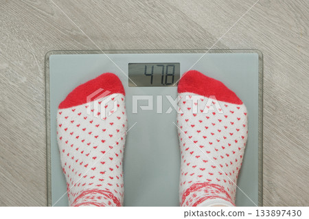 Feet in Cozy Christmas Socks Standing on Digital Bathroom Scales, New Year's Resolution Concept. Feet in Cozy Christmas Socks Standing on Digital Bathroom Scales, New Year's Resolution Concept. 133897430