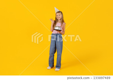 Preteen girl playfully posing with birthday cake while wearing party hat 133897489