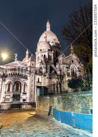 Illuminated Sacre-Coeur Basilica rises above cobblestone street in Montmartre district of Paris, France. Historic church features white stone architecture and Roman-Byzantine style in Ile-de-France 133897504