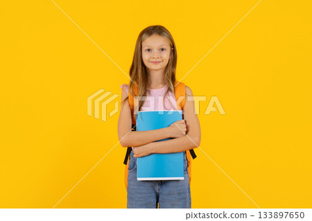 Preteen girl standing with notebook and backpack on bright yellow studio background 133897650