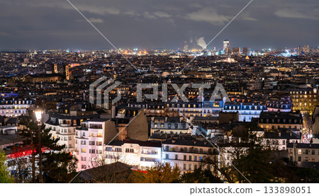 Cityscape of Paris, France glows at night seen from Montmartre hill. Panoramic view features illuminated rooftops, streetlights, and urban horizon under cloudy sky in Ile-de-France region 133898051