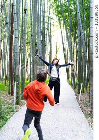 A delighted woman spreads her arms wide in a bamboo forest while a boy runs playfully ahead. This scene captures the essence of family bonding in a tranquil environment. A delighted woman spreads her arms wide in a bamboo forest while a boy runs playfully ahead. This scene captures the essence of family bonding in a tranquil environment. 133899559