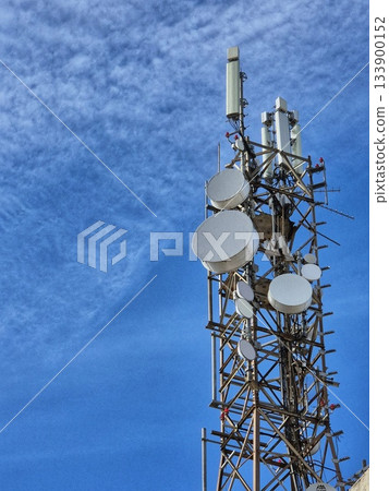 A large communication tower (communication tower) with numerous antennas and satellite dishes, photographed against a bright blue sky. 133900152