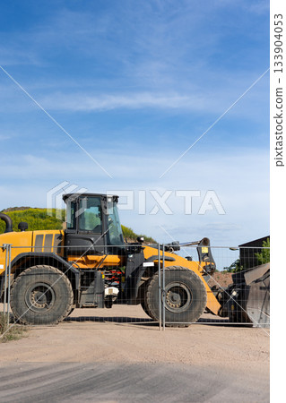 Bright yellow wheel loader stands behind metal fence on dusty construction ground. Sunlight highlights large tires, cabin and bucket while green hill and buildings form calm industrial backdrop 133904053