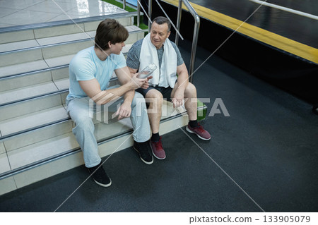 Adult man sitting in gym talking with younger trainer, resting after workout and receiving guidance for improving physical fitness. Senior Man Working Out in Gym Adult man sitting in gym talking with younger trainer, resting after workout and receiving guidance for improving physical fitness. Senior Man Working Out in Gym 133905079