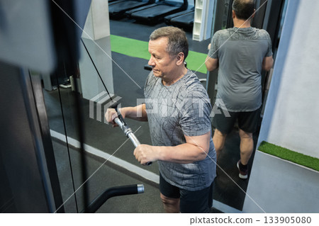 Senior man presses cable handle downward while training triceps in gym, maintaining controlled movement and strength focus. Working Out 133905080