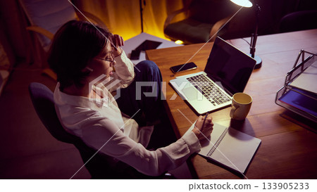 Woman working late and writing notes while reviewing laptop information. Woman working late and writing notes while reviewing laptop information. 133905233