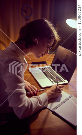 Woman writing notes near laptop with wireless earbuds during study session. Woman writing notes near laptop with wireless earbuds during study session. 133905234