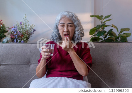 Elderly Woman Taking Pill with Water on Sofa Elderly Woman Taking Pill with Water on Sofa 133906203