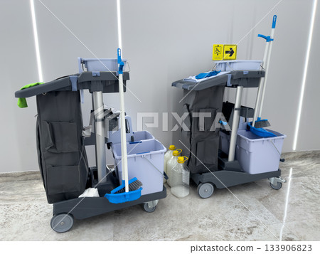 Two cleaning carts in grey hallway. Floor maintenance and cleanliness in public building. Cleaning carts with mops and water buckets. Cleanliness and getting rid of dirt with professional equipment Two cleaning carts in grey hallway. Floor maintenance and cleanliness in public building. Cleaning carts with mops and water buckets. Cleanliness and getting rid of dirt with professional equipment 133906823