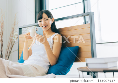 一位年輕女子躺在床上喝咖啡，同時看著她的智慧型手機。 133907537