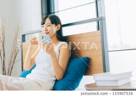 一位年輕女子躺在床上喝咖啡，同時看著她的智慧型手機。 133907538