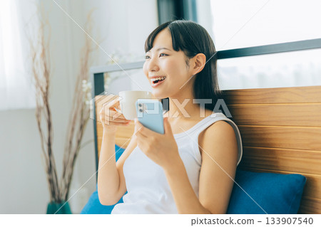 一位年輕女子躺在床上喝咖啡，同時看著她的智慧型手機。 133907540