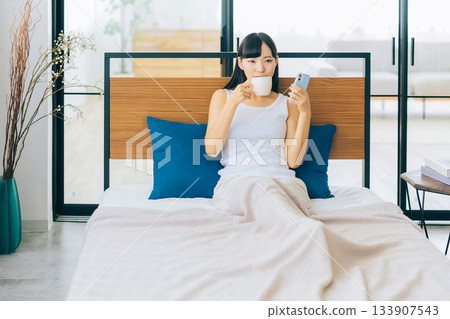 一位年輕女子躺在床上喝咖啡，同時看著她的智慧型手機。 133907543