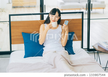 一位年輕女子躺在床上喝咖啡，同時看著她的智慧型手機。 133907544