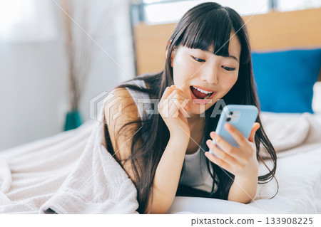 一位女士在床上開心地看著她的智慧型手機。 133908225