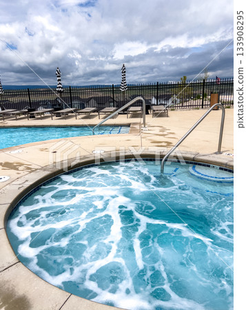 A hot tub filled with foamy water is featured in a spa area with distant views. Lounge chairs, railings, and a wide open sky contribute to a relaxing atmosphere. 133908295