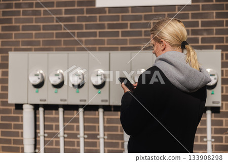 Middle-aged woman checking multiple outdoor electricity meters and entering readings on her smartphone beside a brick building Middle-aged woman checking multiple outdoor electricity meters and entering readings on her smartphone beside a brick building 133908298