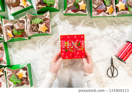 A festive top-down shot of hands wrapping a Christmas cookie gift box, surrounded by neatly arranged holiday treat boxes. The scene showcases decorated gingerbread cookies, festive packaging, and 133908313