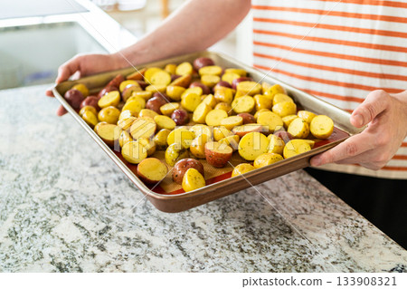 In a modern kitchen, an array of halved, multicolored marble potatoes are arranged on a baking pan lined with a silicone liner. The roasting process infuses the kitchen with a mouthwatering aroma In a modern kitchen, an array of halved, multicolored marble potatoes are arranged on a baking pan lined with a silicone liner. The roasting process infuses the kitchen with a mouthwatering aroma 133908321
