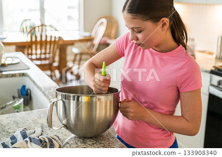 A girl in a pink shirt is focused on mixing cookie dough in a large metal bowl. The kitchen around her is warmly lit with granite countertops and wood furniture. A girl in a pink shirt is focused on mixing cookie dough in a large metal bowl. The kitchen around her is warmly lit with granite countertops and wood furniture. 133908340