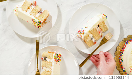 Two slices of vanilla cake with rainbow sprinkles and white frosting are served on round white plates. The scene is styled with gold forks on a marble surface, evoking a festive and cheerful mood. Two slices of vanilla cake with rainbow sprinkles and white frosting are served on round white plates. The scene is styled with gold forks on a marble surface, evoking a festive and cheerful mood. 133908344