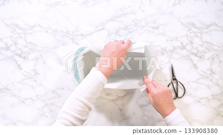 A person places parchment paper into a blue ceramic baking dish on a marble surface. Scissors are nearby for trimming excess paper. A person places parchment paper into a blue ceramic baking dish on a marble surface. Scissors are nearby for trimming excess paper. 133908380