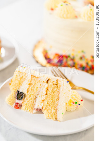 A vanilla cake is cut open with a golden spatula, revealing fruit-studded layers and piped frosting. The exposed interior shows soft sponge and cream detail. 133908390