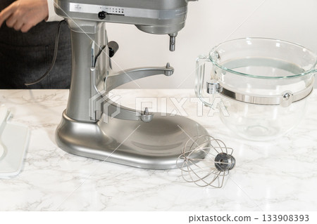 A stand mixer is set up with a glass mixing bowl containing butter, ready to begin making lemon spritz cookies. The wire whisk attachment is prepared for the mixing process. 133908393