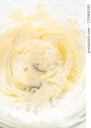 A close-up view of smooth and airy cream cheese filling whipped to perfection. The soft texture and rich color are highlighted in the glass mixing bowl, showcasing the ideal consistency for spreading 133908395