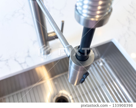 A modern kitchen faucet with a pull-down sprayer head positioned above a ridged stainless steel sink. The clean design is enhanced by a bright countertop and subtle reflections. 133908398