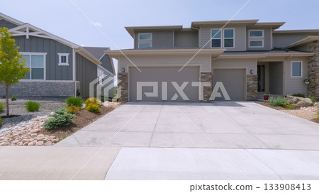A modern suburban house features a two-car garage, stone trim, and a minimalistic landscaped front yard. The driveway is clean and wide under a clear blue sky. 133908413