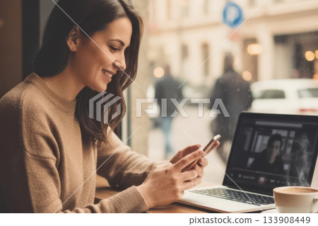 A smiling woman sits at a cafe table using her smartphone with a laptop and cup of coffee nearby. The daylight streaming through the window creates a pleasant and productive atmosphere for social A smiling woman sits at a cafe table using her smartphone with a laptop and cup of coffee nearby. The daylight streaming through the window creates a pleasant and productive atmosphere for social 133908449