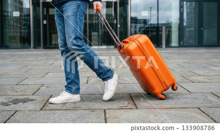 Individual walking with bright orange suitcase on pavement, showcasing travel lifestyle and modern urban environment with stylish footwear Individual walking with bright orange suitcase on pavement, showcasing travel lifestyle and modern urban environment with stylish footwear 133908786